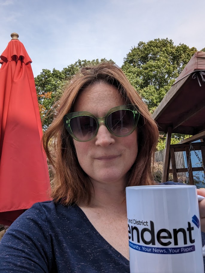 A woman wearing a blue t-shirt and green sunglasses, holding a mug with Caterham Independent written on it. She's standing in a garden, in front of a red parasol and a wooden swing seat