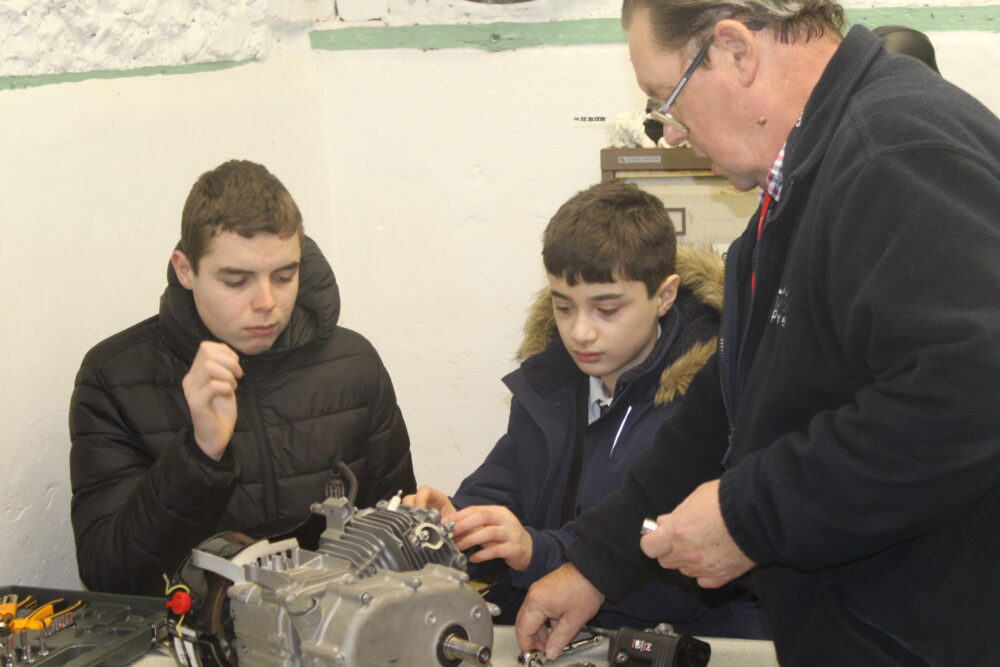 An instructor talks to two students looking at an engine