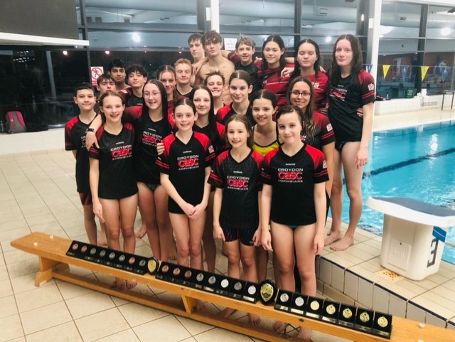 A swimming club, standing in front of a pool. A bench of medals is in front of them