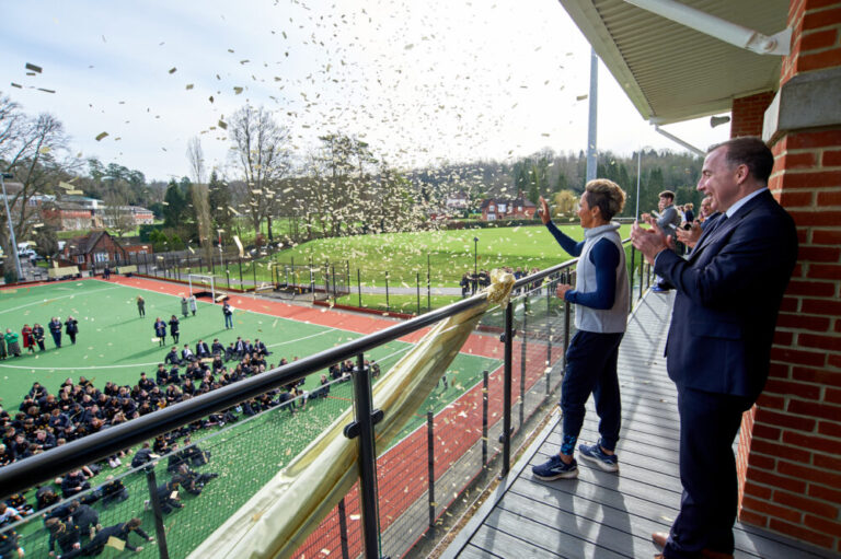 Dame Kelly Holmes standing on a balcony, with one hand in the air. A man in a suit is standing slightly behind to her left. Below, a large group of students and teaching staff are standing on an Astro turf pitch