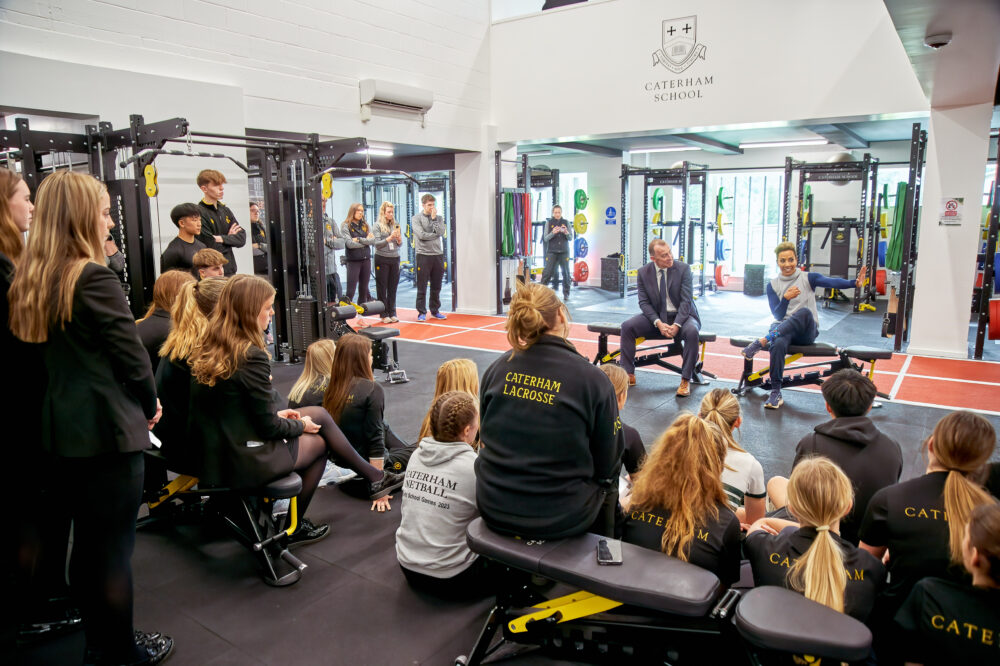 Dame Kelly Holmes sits in front of a group of students in a gym. A man sits to her right