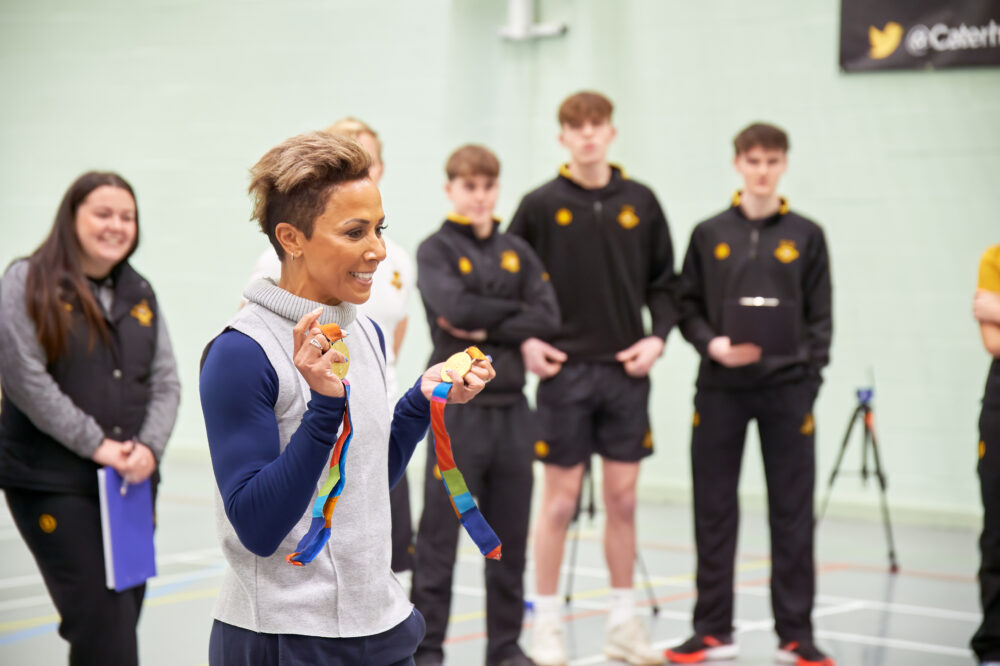 Dame Kelly Holmes stands with her back to four students in sports gear, holding her two Olympic gold medals